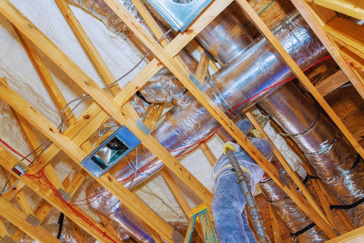 Professional in protective gear spraying insulation around extensive silver insulated duct work in a home attic during HVAC installation or upgrade.