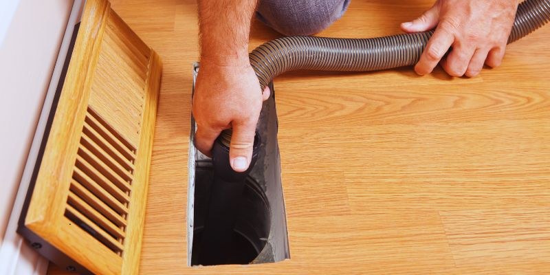 Technician inserting a large vacuum hose into an open floor vent to clean residential duct work and improve indoor air quality.