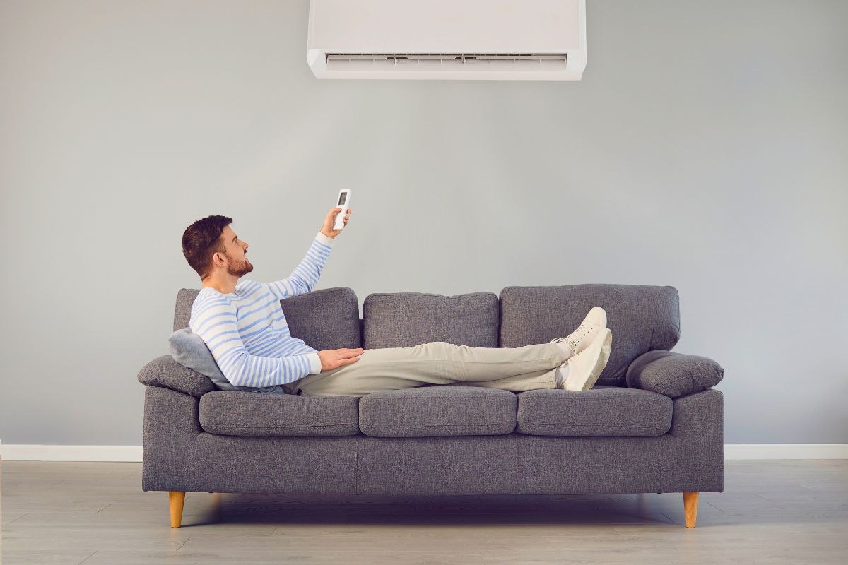 Man relaxing on the sofa pointing remote at AC unit, frustrated because the house feels humid with ac running