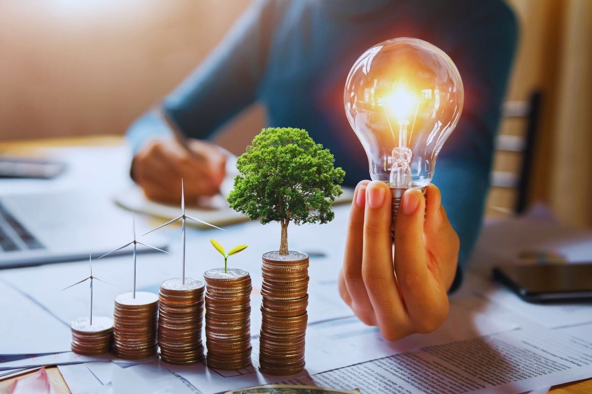 A person holding a glowing light bulb above stacked coins with wind turbines and trees in the background, illustrating what is energy conservation and how smart energy use saves money and protects the environment.