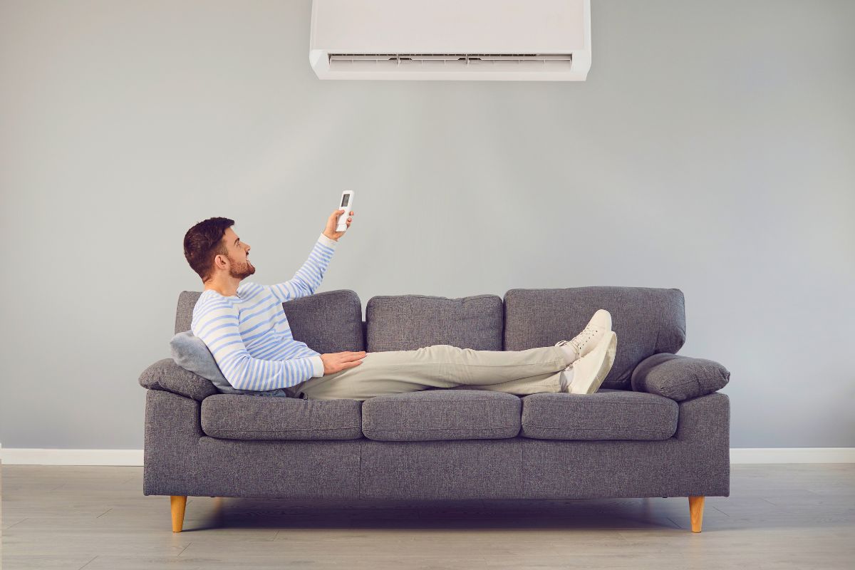 Man relaxing on the sofa pointing remote at AC unit, frustrated because the house feels humid with ac running
