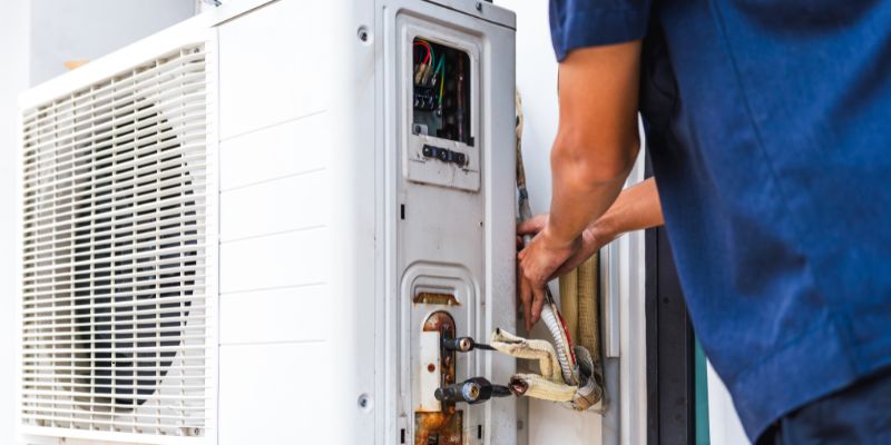 Professional HVAC technician in blue uniform performing hvac rough in on an outdoor condenser unit, accessing the electrical panel and connecting refrigerant lines during installation.