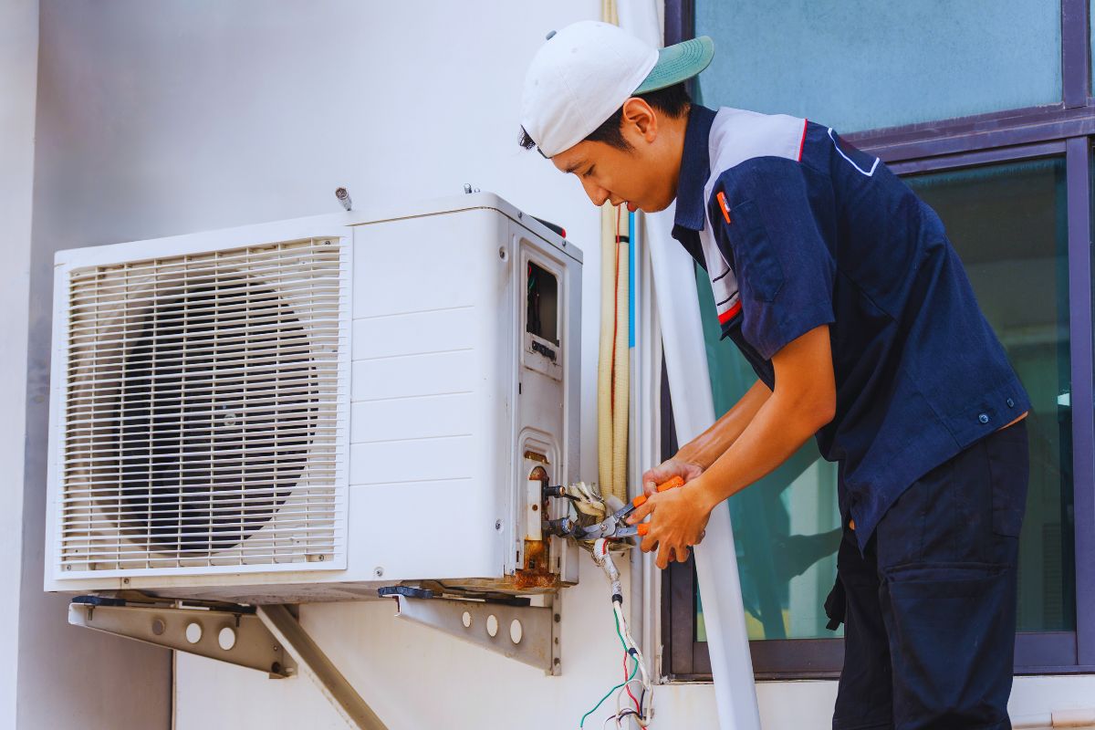 Professional HVAC technician in uniform servicing an outdoor air conditioning condenser unit, using pliers to work on wiring and components on the side of a building, demonstrating the mechanical and refrigerant system involved when learning how does an air conditioner work