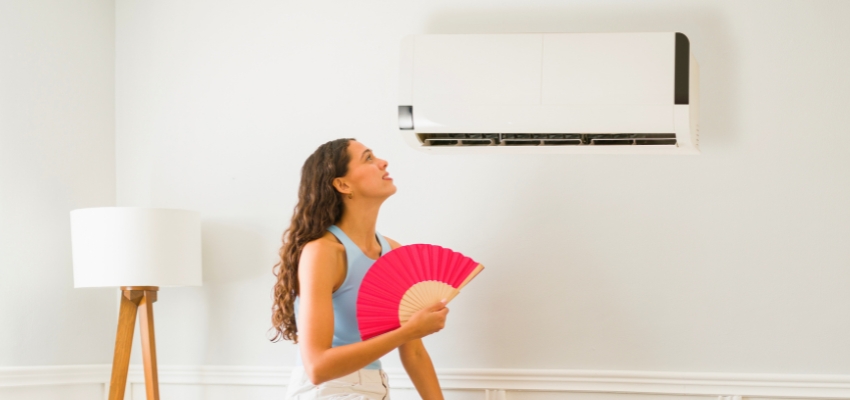 Woman looking up at a wall-mounted air conditioner, illustrating the importance of a properly functioning AC capacitor for cooling comfort.