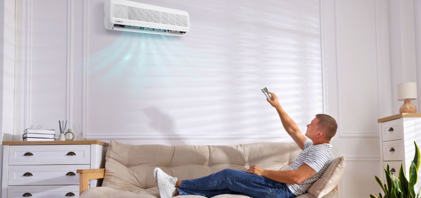 Man relaxing on sofa using remote to control wall-mounted AC, illustrating common issues when your ac making noise