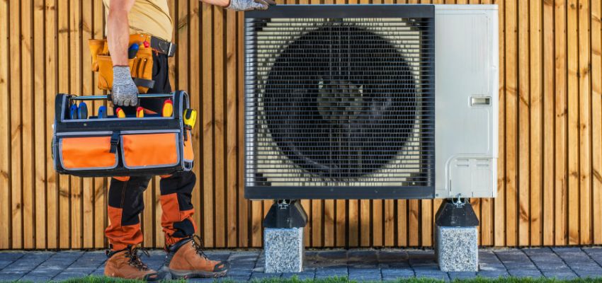 Technician with toolbox standing beside a residential outdoor unit during an HVAC tune up.
