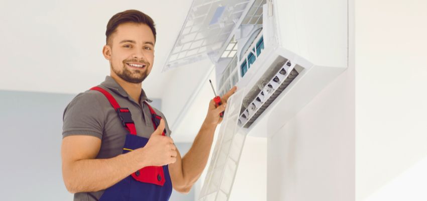 Smiling technician giving a thumbs up during an HVAC tune up while servicing an indoor air conditioning unit.